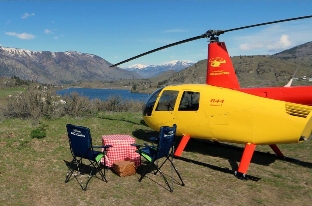A yellow colors Lake Chelan helicopter sitting on a grassy spot overlooking the mountains